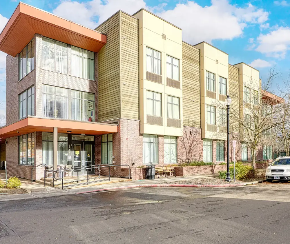 Modern multi-story building with large windows, featuring a ramp at the entrance and parked cars along the street under a sunny sky Modern multi-story building with large windows, featuring a ramp at the entrance and parked cars along the street under a sunny sky
